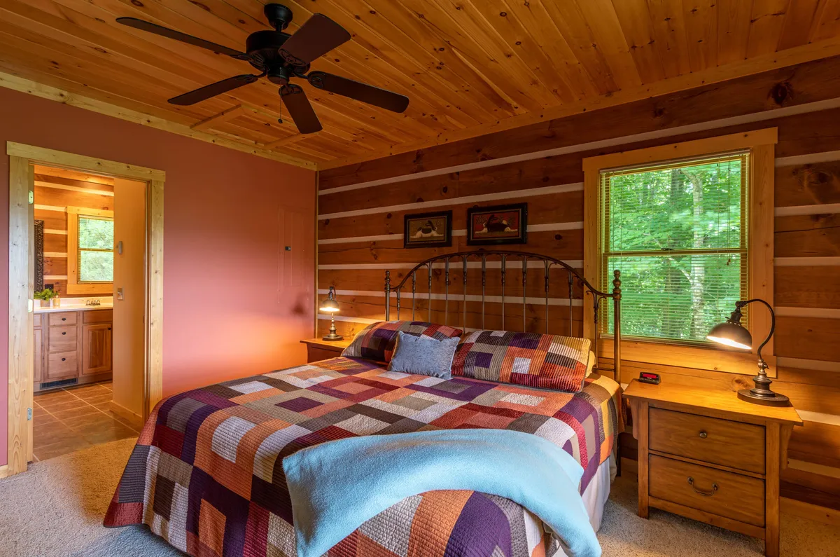 A cozy bedroom featuring a bed with a patterned quilt, wooden ceiling, nightstand with lamps, and a window overlooking trees. The intimate size and soft lighting create a peaceful, private atmosphere perfect for resting.