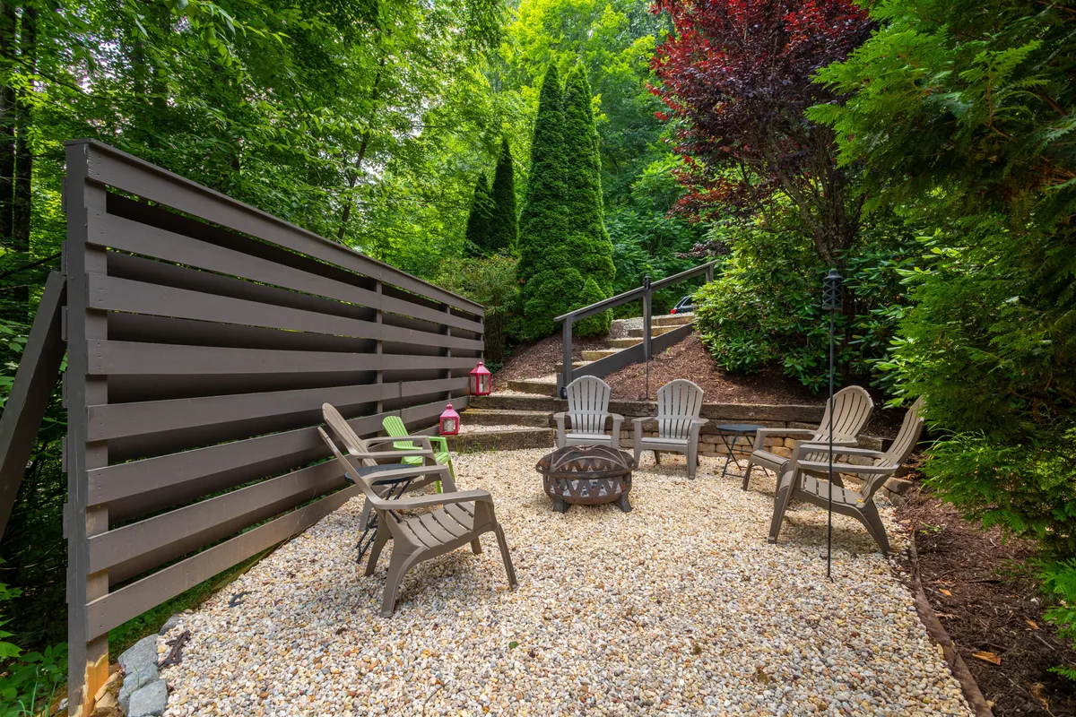 An inviting exterior scene featuring a stone-ringed fire pit surrounded by Adirondack-style chairs, set amid lush greenery. The area is ideal for evening gatherings, storytelling, and making s'mores under an open sky, evoking a sense of family togetherness and classic outdoor fun.
