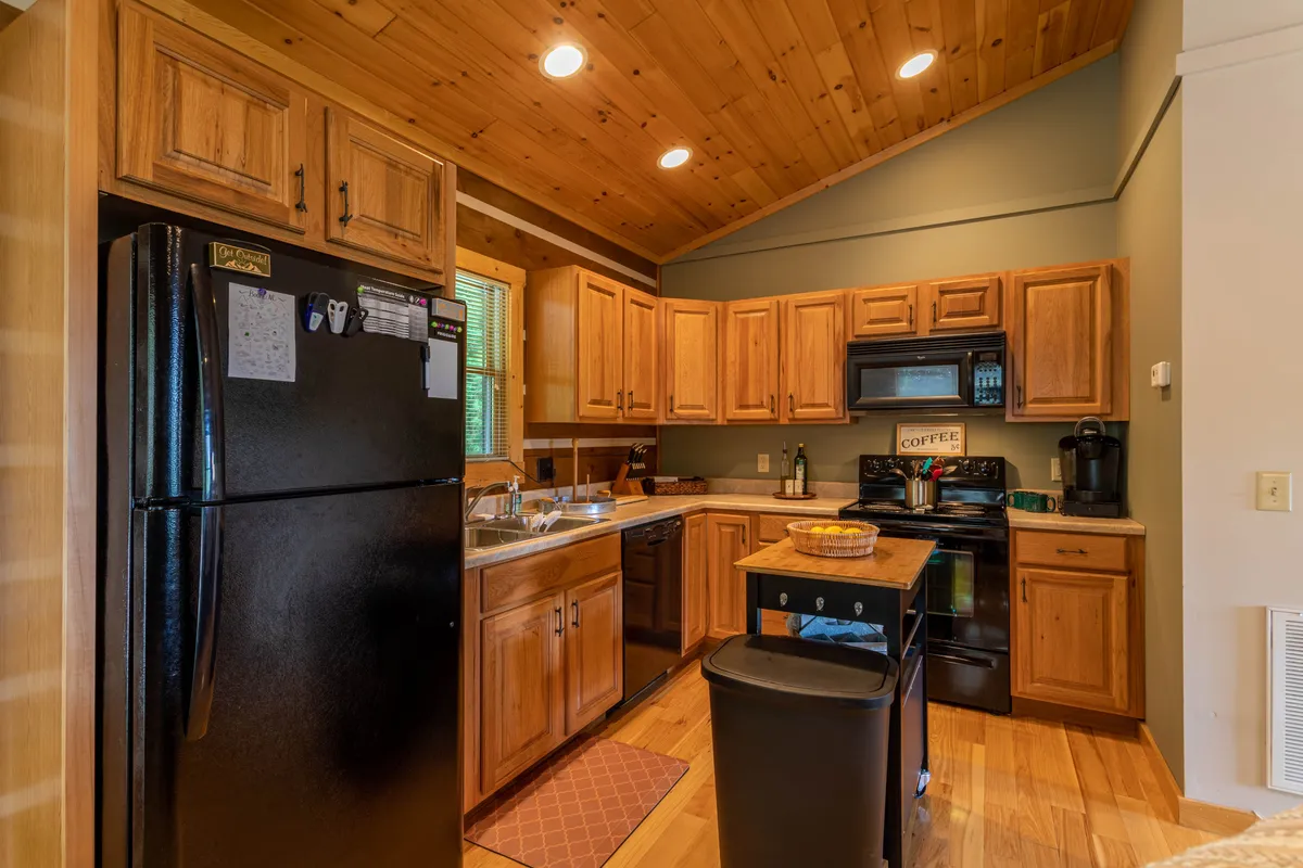 A cozy kitchen scene with wooden cabinets, a black refrigerator, kitchen island, coffee maker, and sink. The space invites collaborative cooking and socializing, with ample room for preparing meals and brewing coffee together.