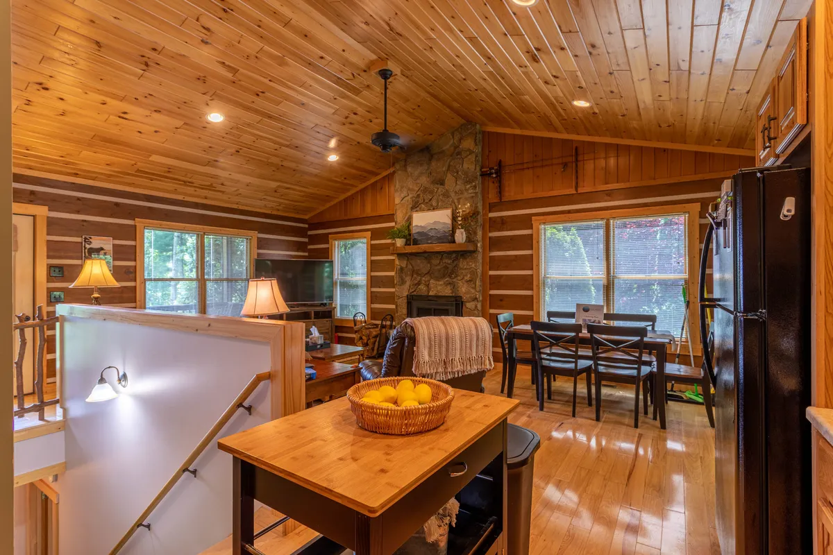 A wide view of an open-plan living room and kitchen, featuring a rustic stone fireplace, wooden ceiling, dining area, and a central kitchen island adorned with a fruit bowl. Comfortable seating is grouped around the fireplace, while large windows let in natural light, emphasizing the inviting, communal space.