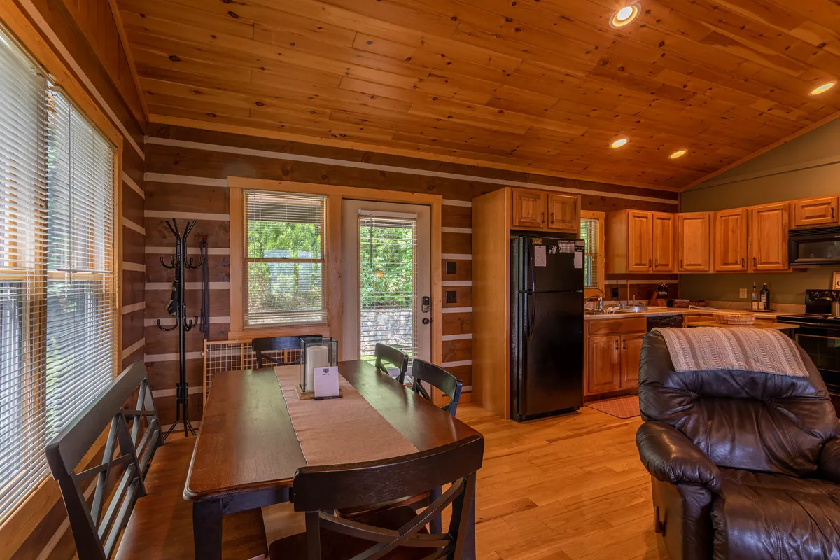 An open dining and kitchen area with a wooden dining table, recliner chair, and large windows that let in plenty of light. The space is set up for shared meals, relaxed conversation, and easy supervision of children or pets. The warm, uncluttered design suggests practical comfort for families.