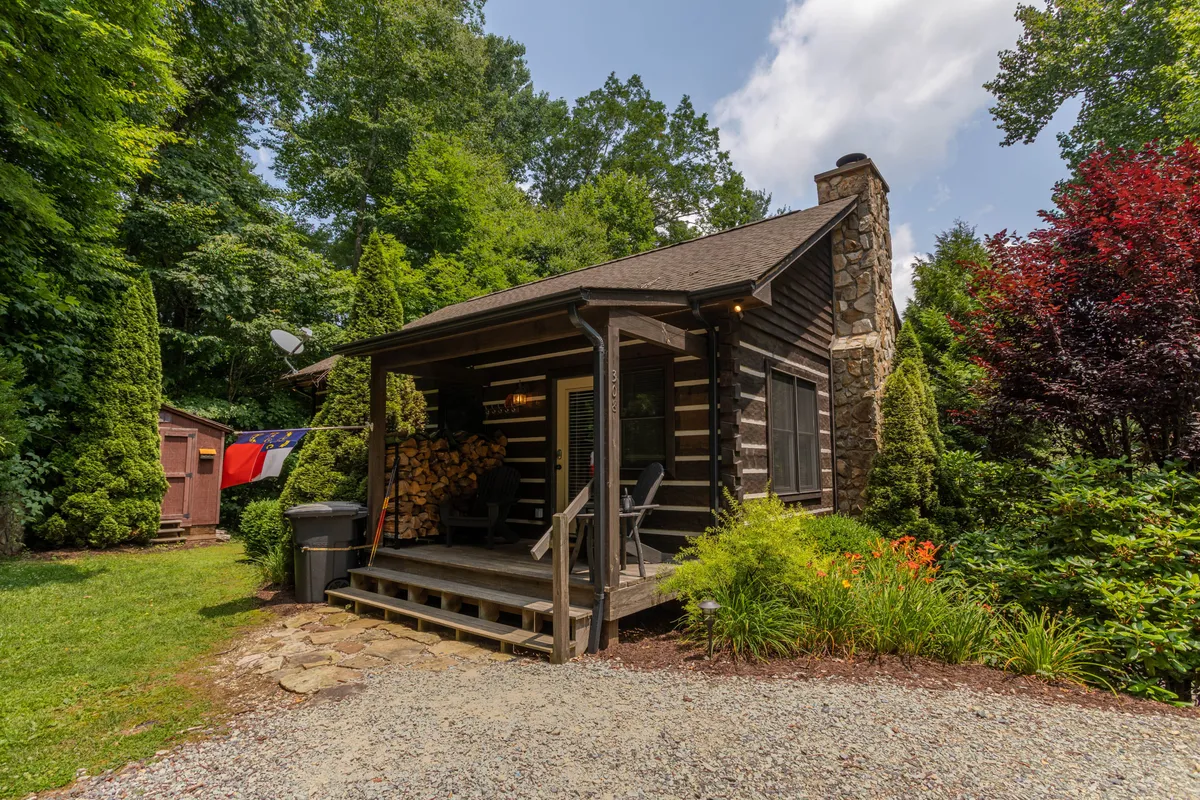 The hero image showcases a picturesque log cabin nestled in lush greenery, with a sturdy stone chimney, a tidy woodpile stacked on the porch, and vibrant flower beds and shrubbery adding pops of color. The inviting porch hints at leisurely mornings with coffee, while the surrounding natural beauty suggests peaceful relaxation and evenings by the fire pit.