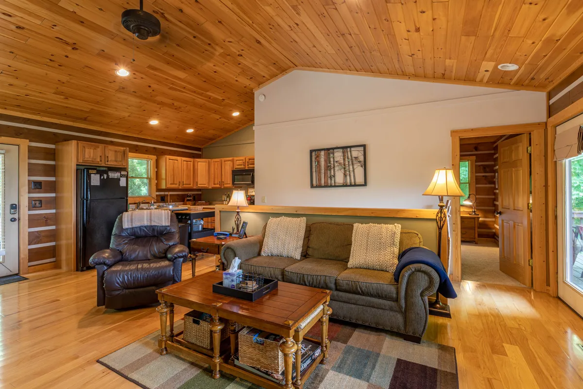 A welcoming living area with a comfortable sofa, coffee table, lamp, and wooden ceiling, seamlessly blending into the kitchen area. The space is designed for both relaxation and socializing, with plenty of room for everyone to gather.