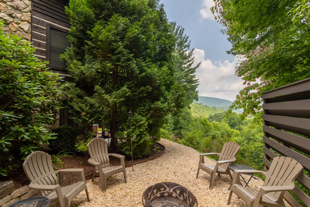 A cozy fire pit area with wooden chairs sits nestled among lush greenery, with expansive mountain views in the background. The setting is ideal for evening gatherings, stargazing, or sharing s’mores with friends and family.