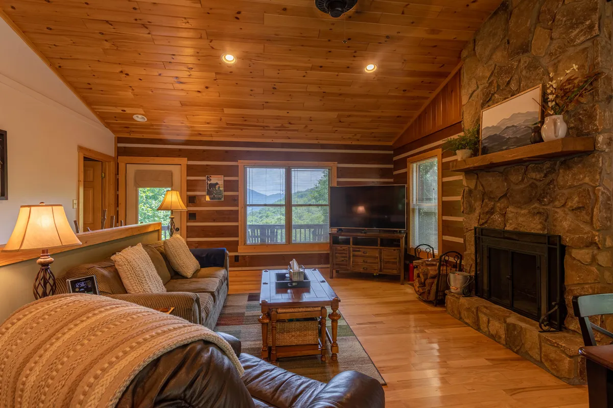 A cozy living room scene featuring a rustic stone fireplace, timber ceiling, comfortable seating, and a large window offering expansive views. The warm lighting and arrangement invite guests to relax and enjoy cozy moments with loved ones.