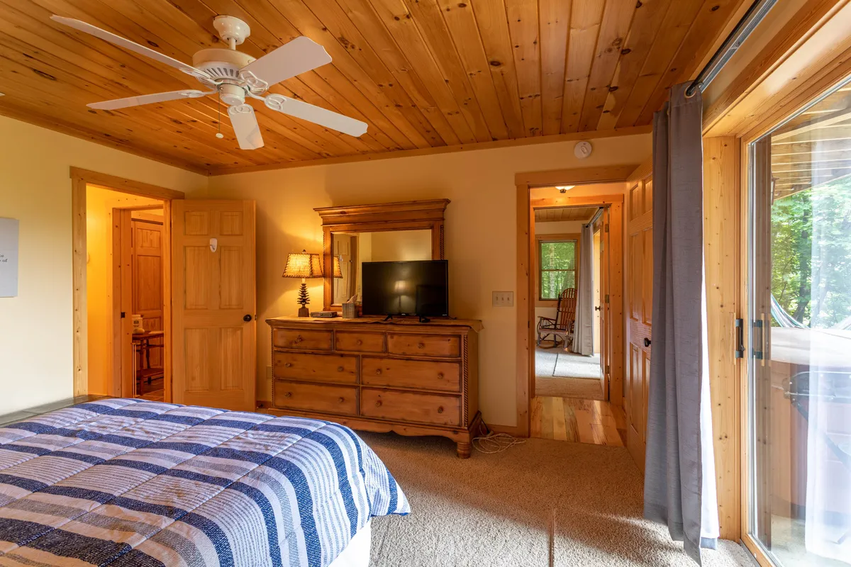 A tranquil bedroom with a plush bed adorned with a blue and white patterned blanket, a wooden dresser with a mirror, a TV atop the dresser, and a sliding glass door leading outside. The space feels airy and bright, with touches that suggest comfort and privacy.