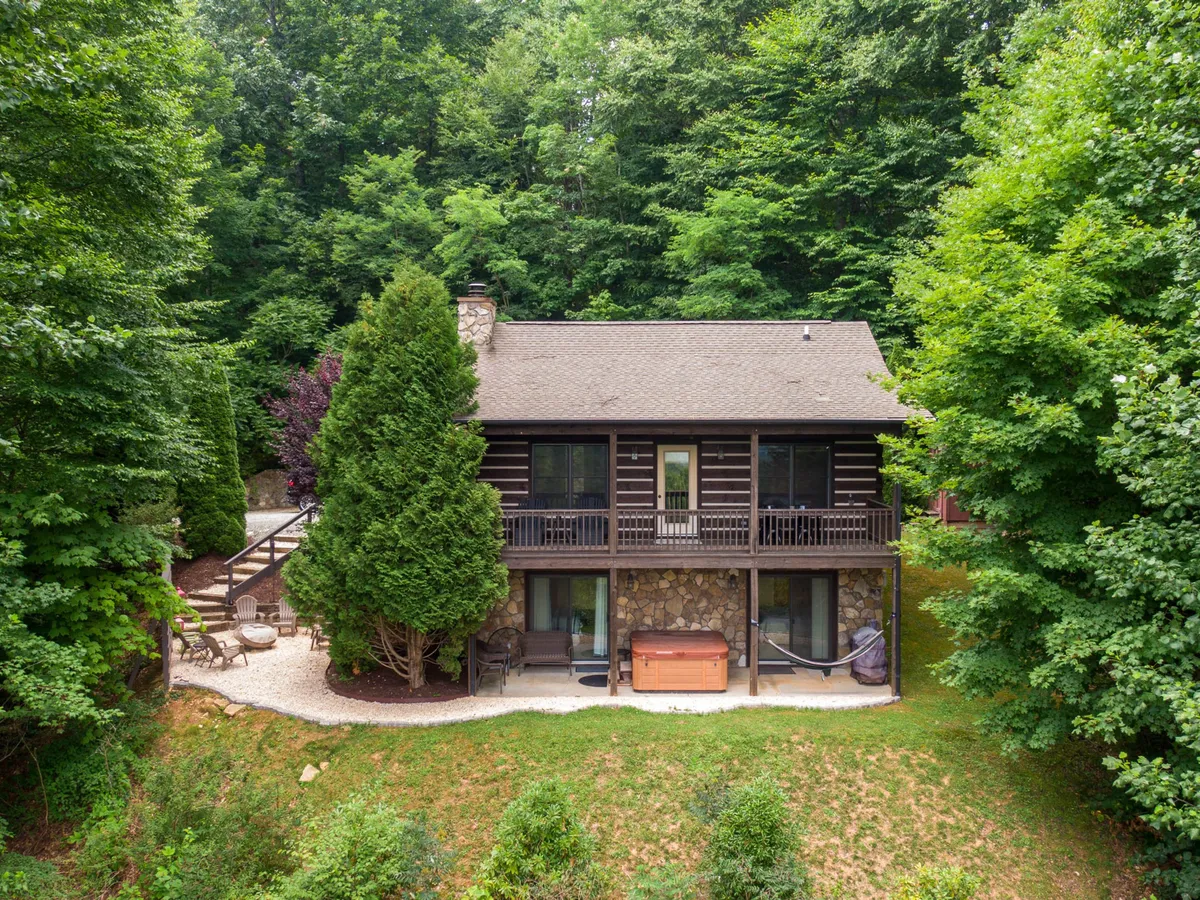 A view of the log cabin's exterior featuring a hot tub, outdoor seating area, and a backdrop of trees. The setting exudes tranquility and offers direct access to nature, while the hot tub on the balcony suggests relaxation after outdoor adventures. The surrounding landscape underscores the mountain getaway appeal.
