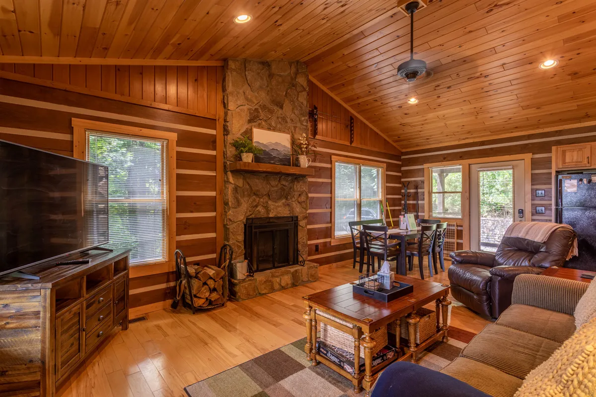 A cozy, spacious living room with a stone fireplace, large sofa, dining table, and television. The room is arranged for relaxation and group activities, suggesting a welcoming environment for families to gather, play games, and watch movies together. Ample natural light adds to the inviting atmosphere.